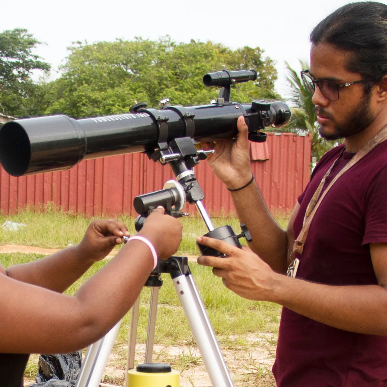 Samir adjusting a refractor telescope on a tripod outdoors while another person steadies the mount