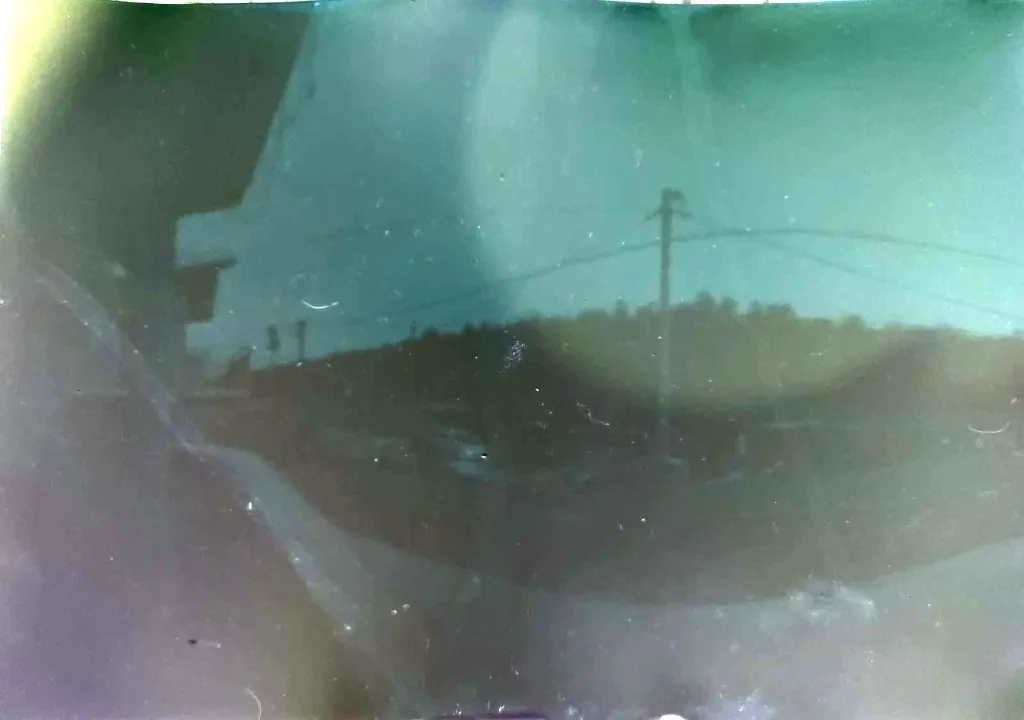 A pinhole solargraph in teal and grey: a roof edge, a utility pole and power lines under a pale sky, with light streaks where the Sun burned into the paper